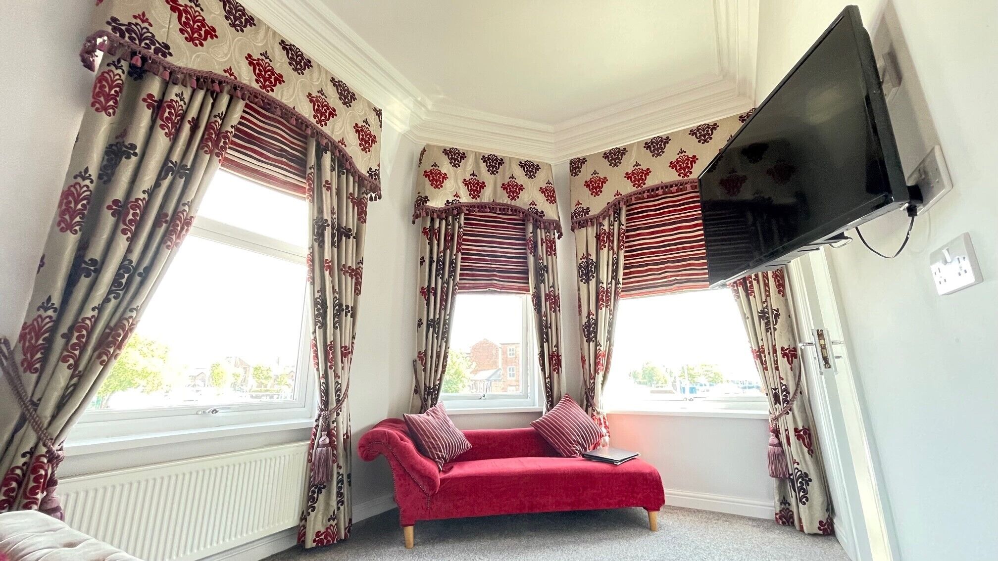 A corner room with a red sofa, patterned curtains, a flat-screen TV, and a radiator.
