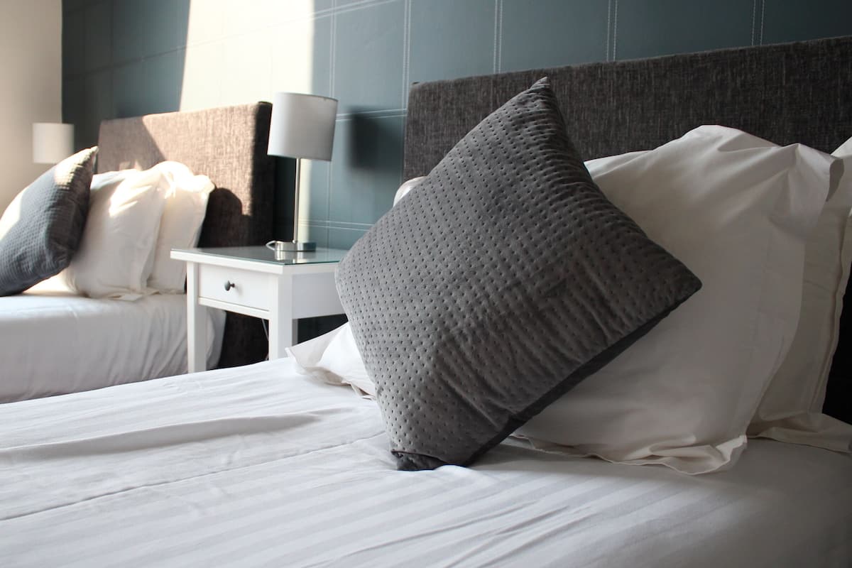 A hotel room with two beds, each with white linens and grey pillows. There is a bedside table with a lamp and a small drawer.