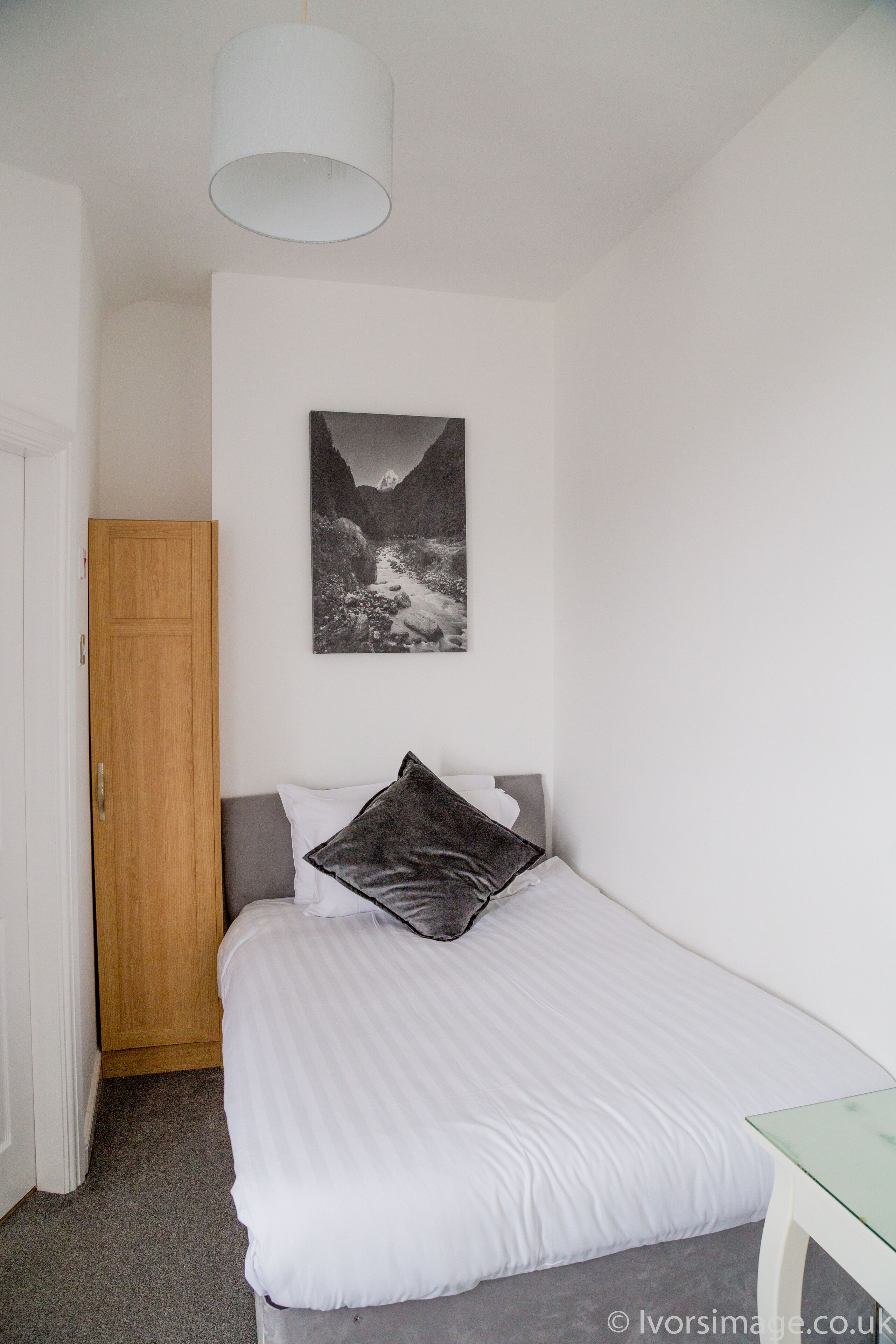A bedroom with a bed, a wooden wardrobe, a painting on the wall, and a small table.