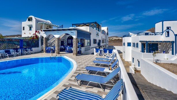 Un área de piscina con sillones de descanso, un edificio de arquitectura blanca y azul, y un cielo azul despejado.