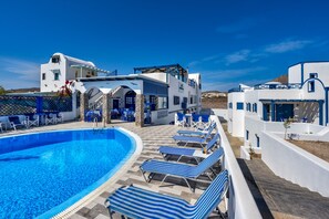 A pool area with lounge chairs, a building with white and blue architecture, and a clear blue sky. - Honeymoon Beach Studios (Santorini)