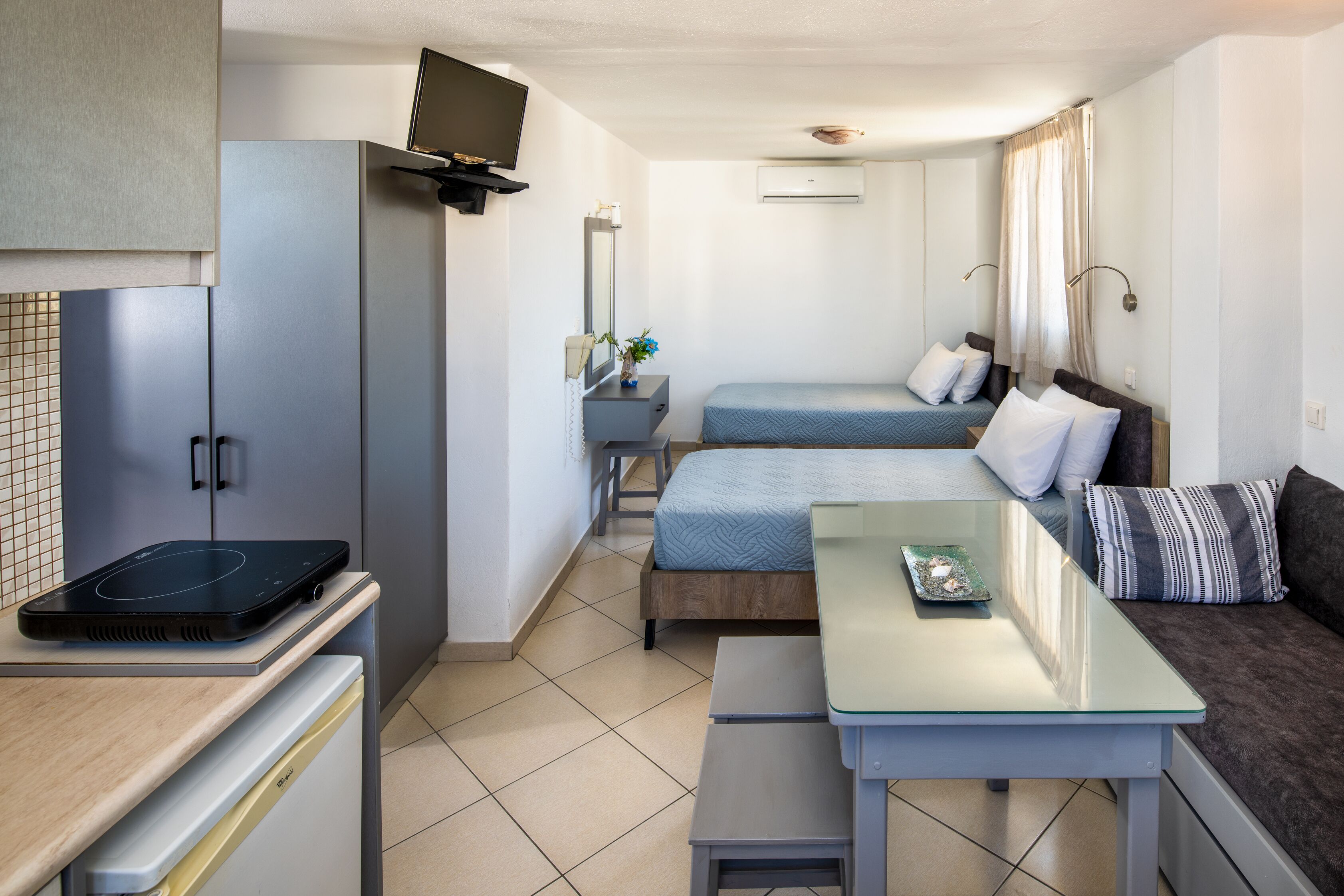 A compact room with a kitchenette, a bed with a blue bedspread, a sofa with striped cushions, and a small table with a tray.