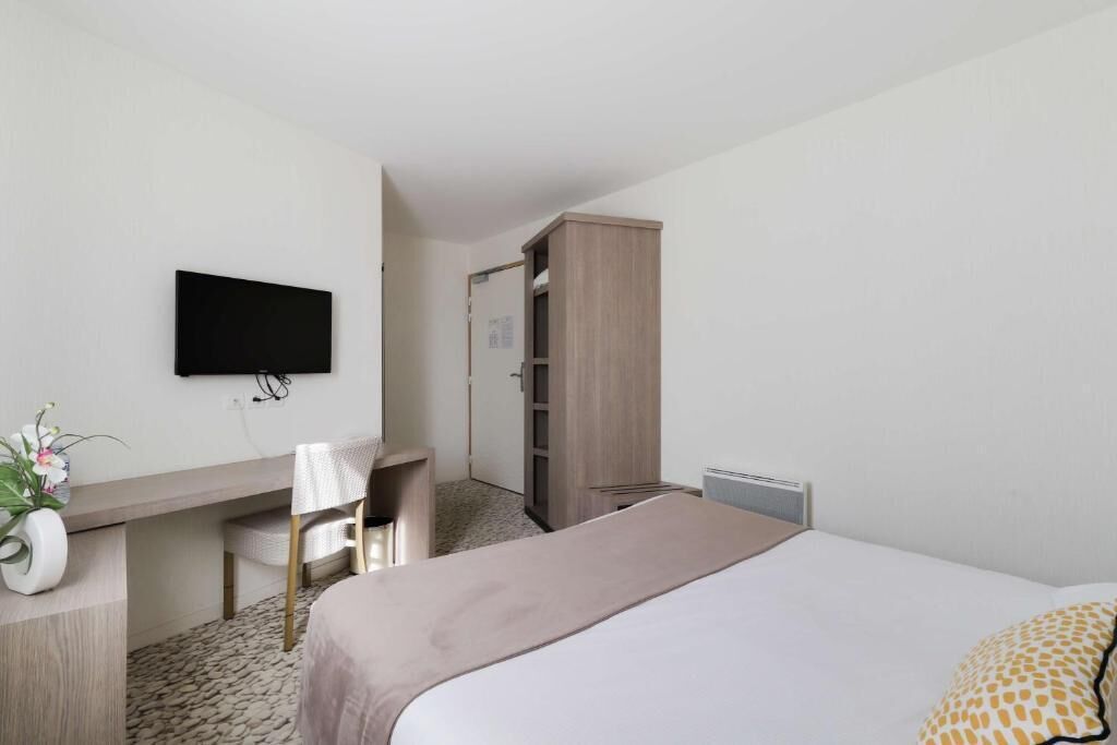 A hotel room with a bed, a desk with a chair, a television mounted on the wall, and a wardrobe.