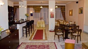 A spacious living area with a dining table, chairs, and a red carpet. There are two chandeliers, a sofa, and a coffee table. - Alexiou Hotel (Kalabaka)