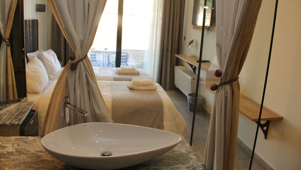 A hotel room with a large oval sink, a bed with white linens, a balcony with a chair, and curtains tied back to reveal the view. - Alexiou Hotel (Kalabaka)