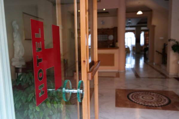A hotel entrance with a glass door, a reception desk, and a patterned rug on the floor. - Alexiou Hotel (Kalabaka)