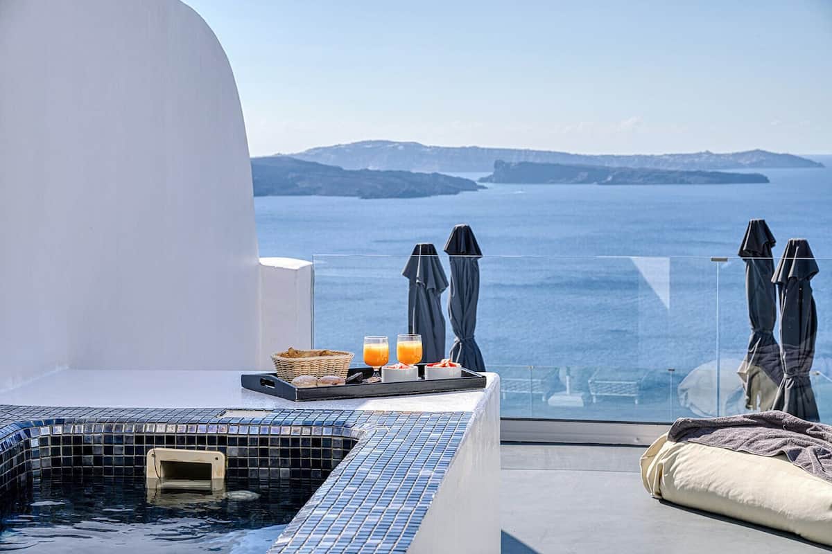 A balcony with a glass balustrade overlooking the sea, a hot tub, and a breakfast tray with orange juice and pastries.