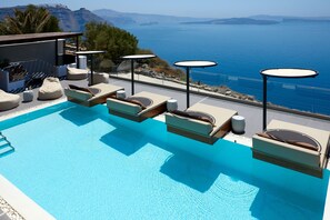 A poolside area with lounge chairs and tables overlooking a body of water. - Oia Suites (Santorini)
