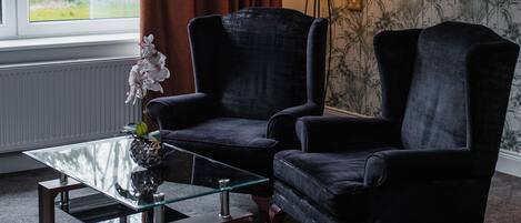 A room with two black armchairs, a glass coffee table, a floor lamp, and a window with curtains.