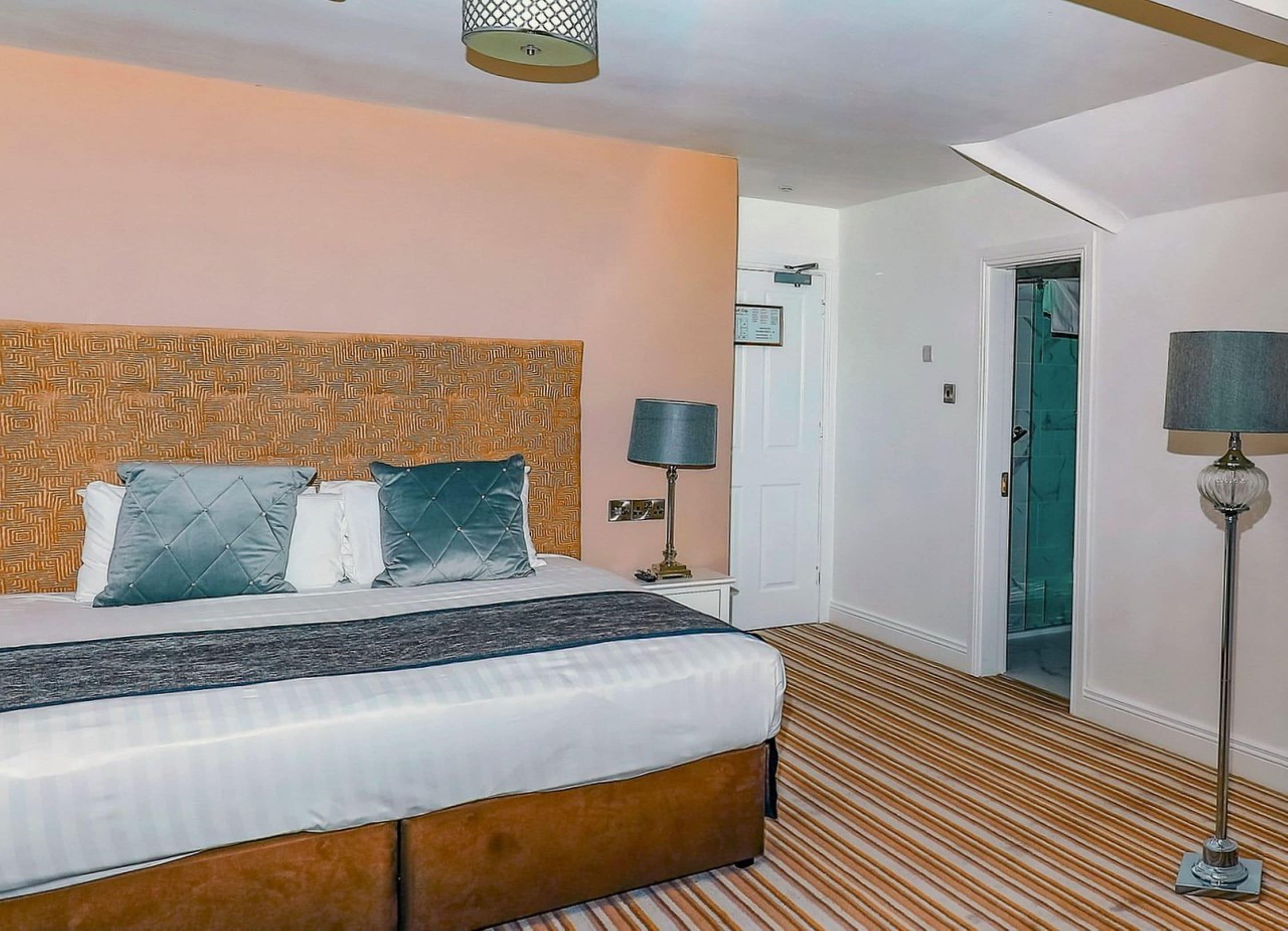 A hotel room with a bed, a nightstand, a lamp, and a door leading to another room.