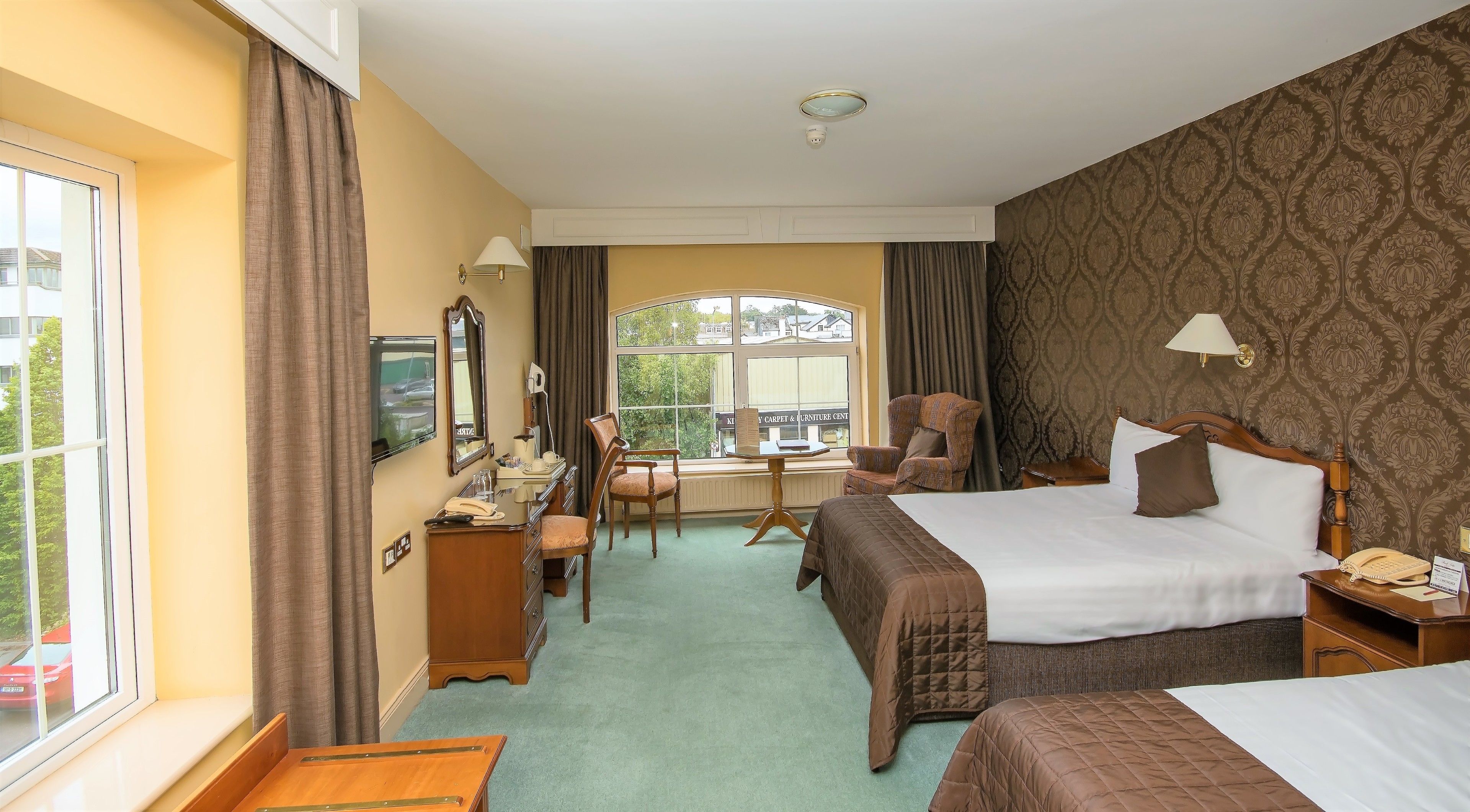 A hotel room with a large bed, a smaller bed, a desk with a chair, a vanity with a mirror, and a window with curtains.