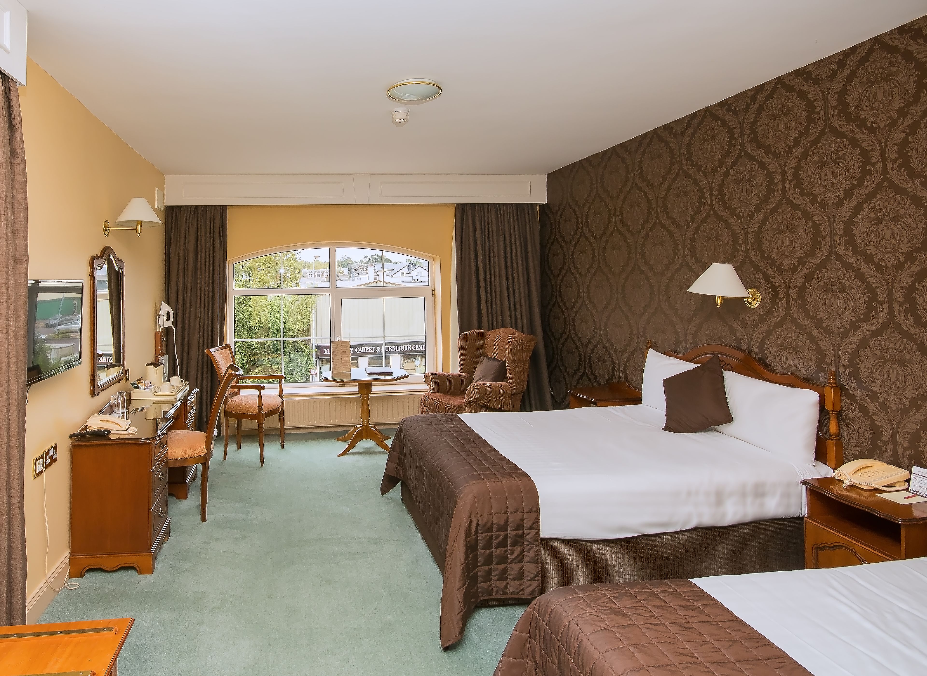 A hotel room with a large bed, two bedside tables, a desk with a chair, a TV, and a window with curtains.
