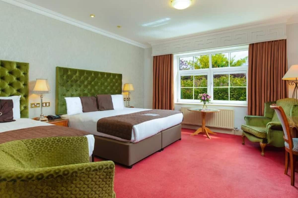 Une chambre d’hôtel avec deux lits, un fauteuil vert, un bureau et une fenêtre avec des rideaux.