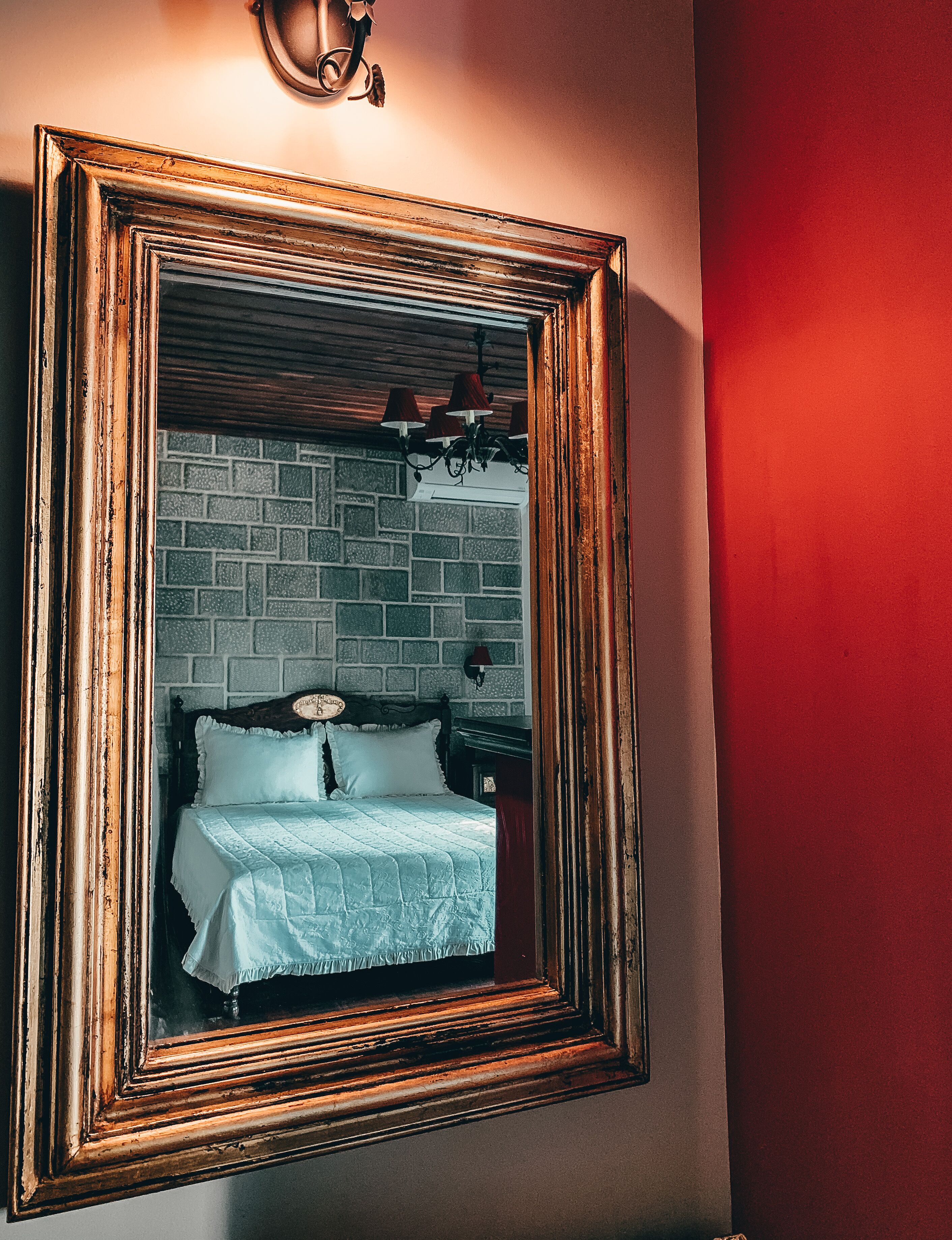 A framed mirror reflecting a room with a brick wall, a bed with white bedding, and a nightstand with a lamp.