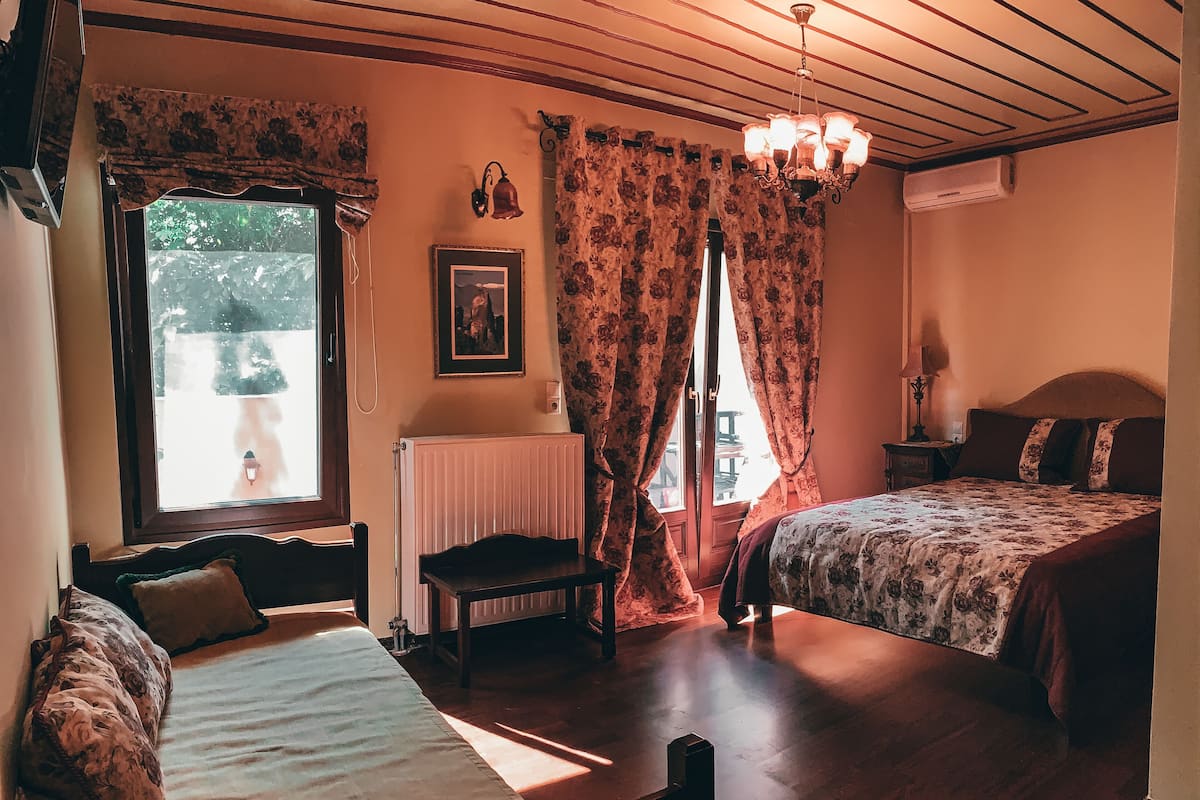 A room with two beds, a radiator, a window with curtains, a chair, a table, and a chandelier.