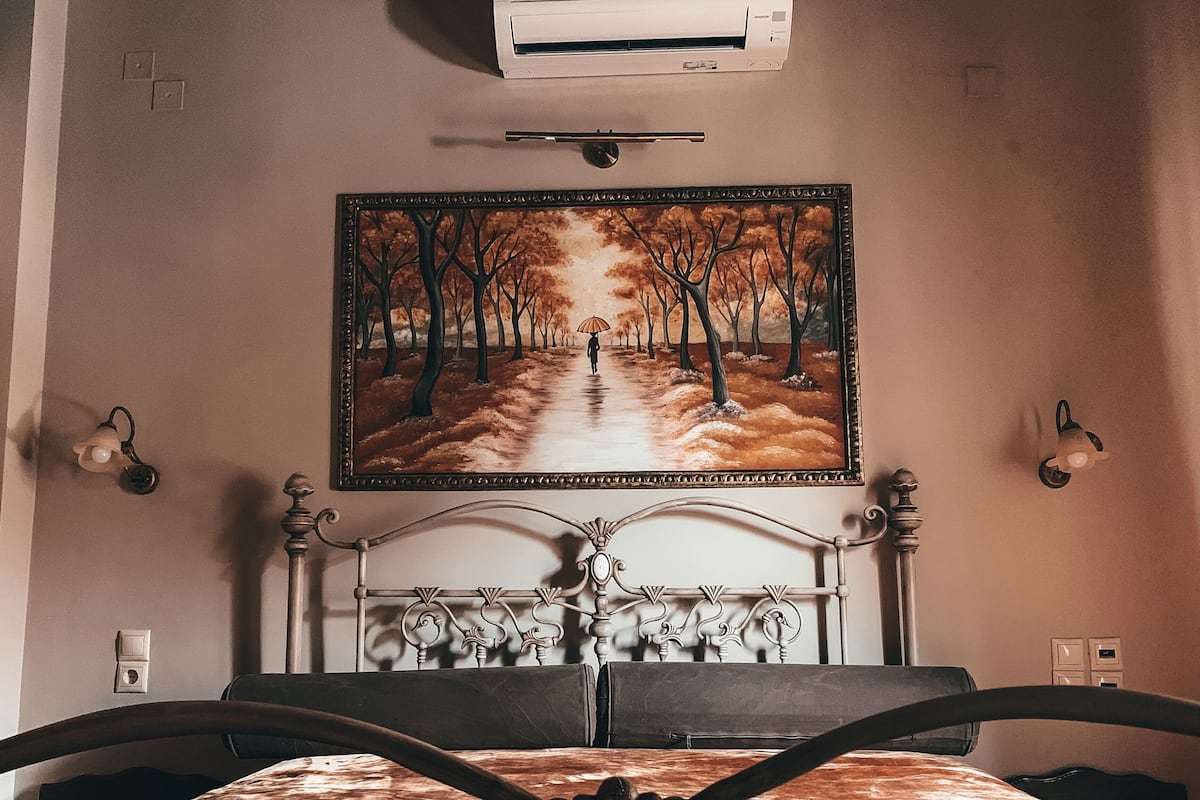 A bedroom with a bed, a headboard, a wall-mounted air conditioner, a painting of a forest path, and wall sconces.