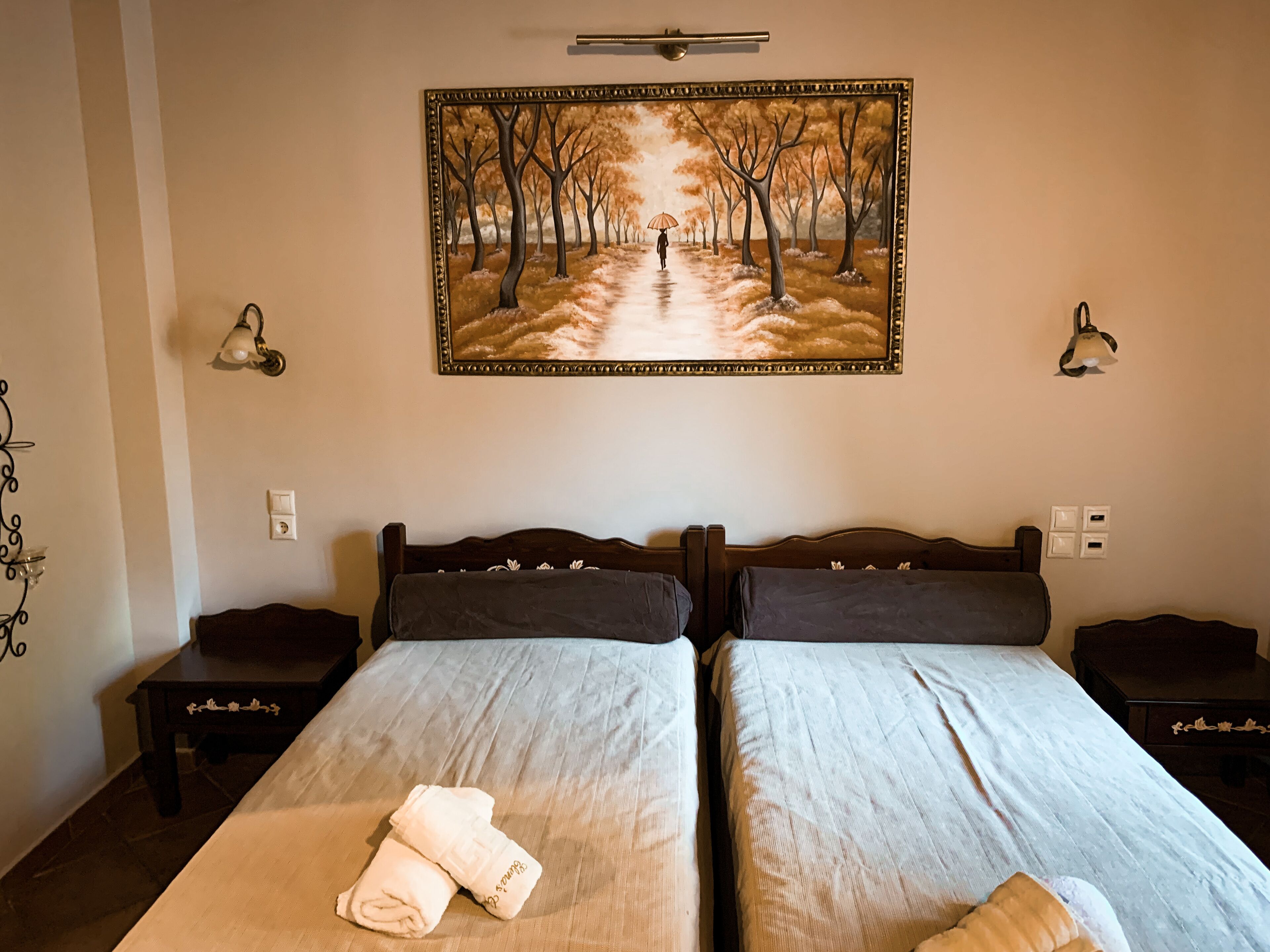 A hotel room with two beds, a painting of a tree-lined path, bedside lamps, and a small table with a lamp.