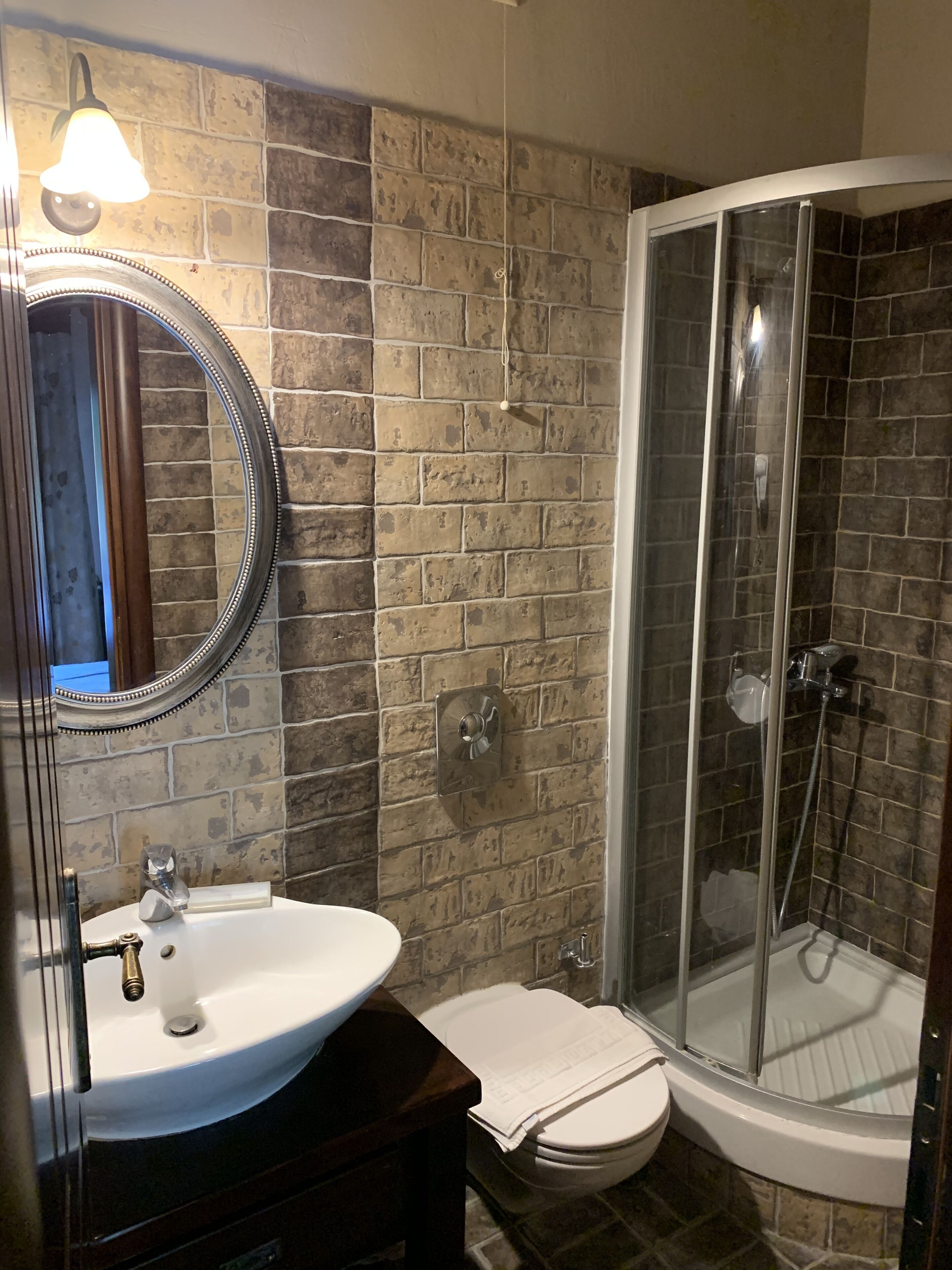 A bathroom with a stone wall, a round mirror, a sink, a toilet, and a glass-enclosed shower.