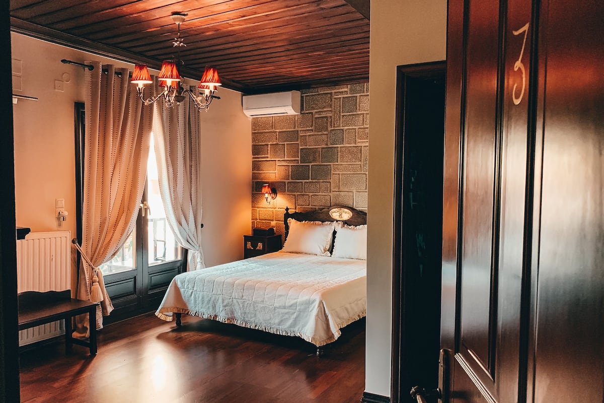 A bedroom with a wooden floor, a bed with a white bedspread, a stone wall, a wooden ceiling, and a chandelier.