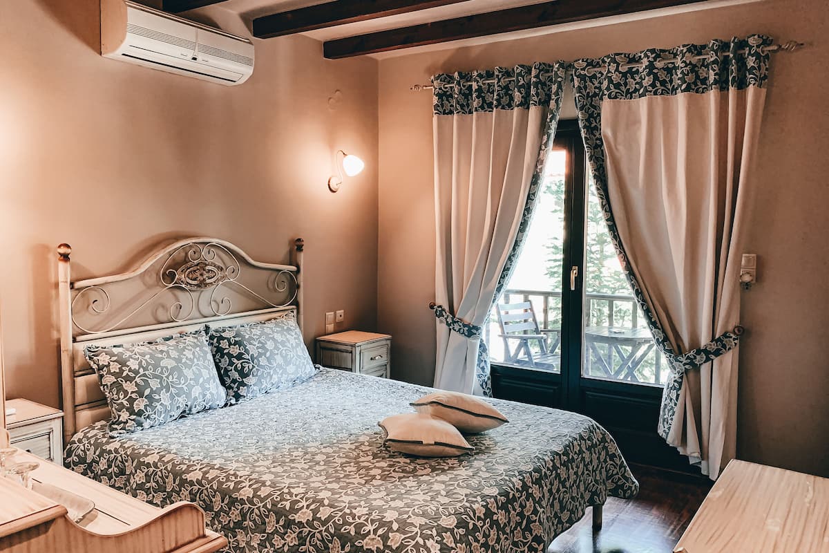 A bedroom with a bed, bedside tables, an air conditioner, a window with curtains, and a view of the outdoors.