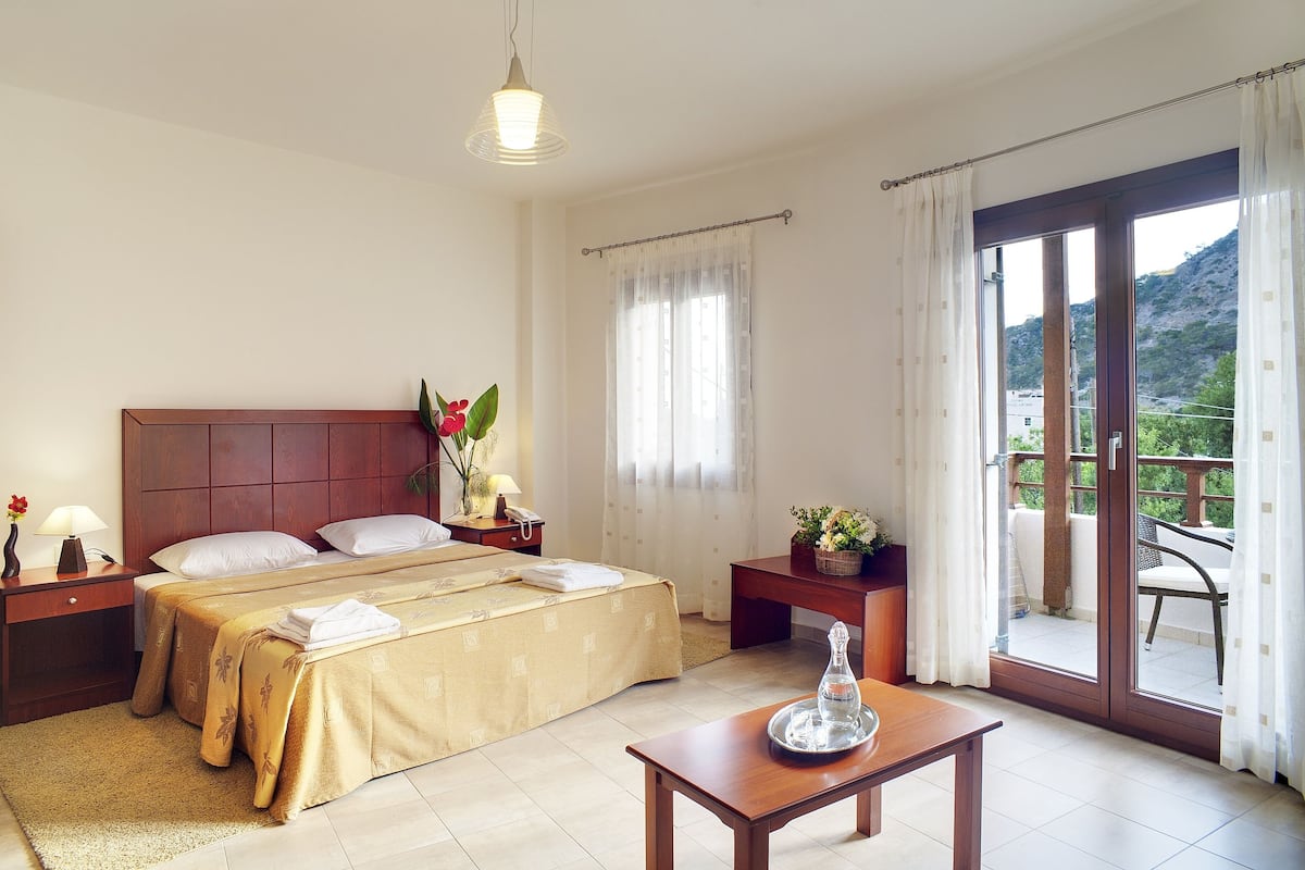 A hotel room with a bed, bedside tables, a nightstand, a lamp, a vase with flowers, a table with a glass pitcher, and a view of a balcony with outdoor furniture.