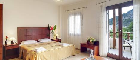 Habitación de hotel con cama, mesitas de noche, lámpara, un florero con flores, una mesa con jarra de vidrio y vista a un balcón con mobiliario exterior.