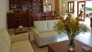 A modern living room with a white sofa, a wooden coffee table, and a vase with flowers. A bar area with shelves of bottles and glasses is visible in the background. - Syia Hotel (Kantanos-Selino)