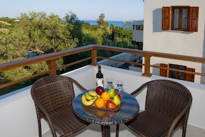 A balcony with wicker chairs, a table with fruit and wine, and a view of the sea and greenery.