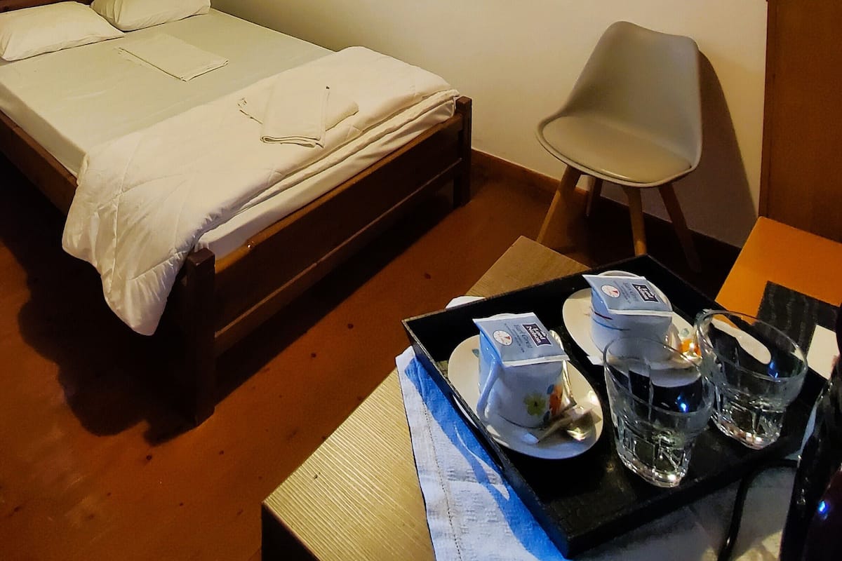 A neatly made bed with a white comforter and pillows, a single chair, a bedside table with a tray of glasses and a bottle, and a hanging lamp.