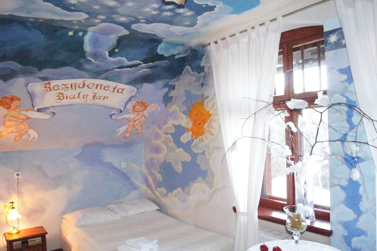 A bedroom with a mural of clouds and stars, a bed, a nightstand, and a window with curtains.