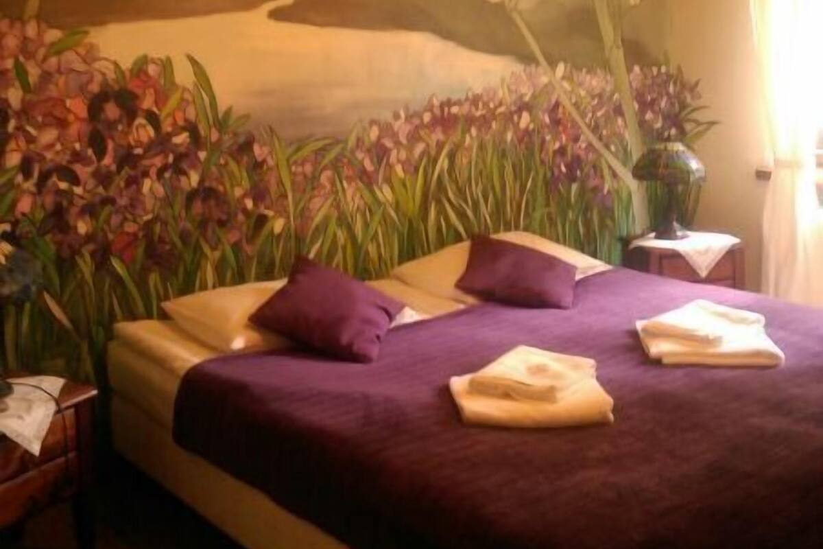 A bedroom with a bed, purple bedding, a nightstand, a lamp, and a wall mural.