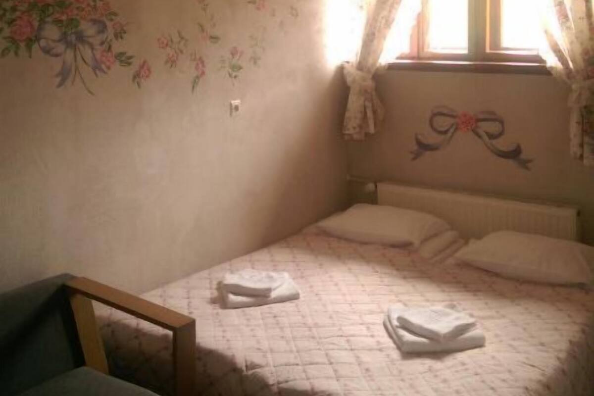 A small, dimly lit room with a single bed, a chair, floral wall decorations, and a window with curtains.