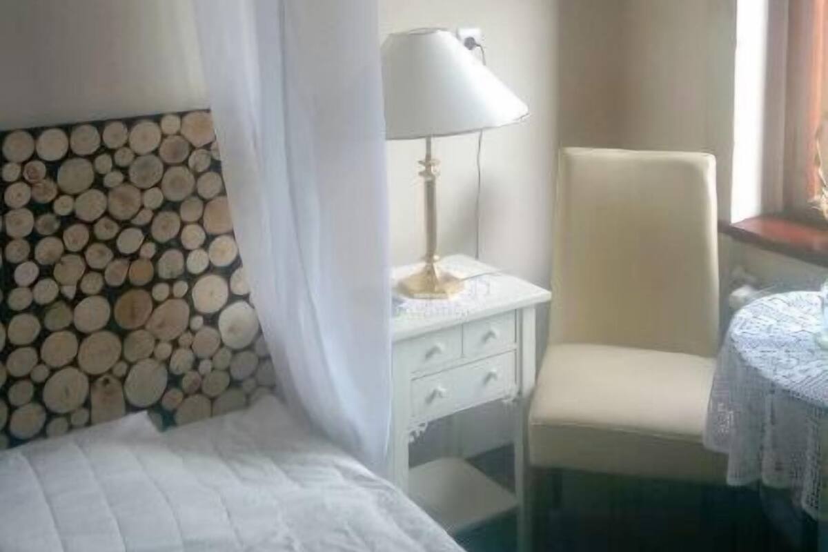 A hotel room with a bed, a chair, a bedside table with a lamp, a wall-mounted painting, and a curtain.