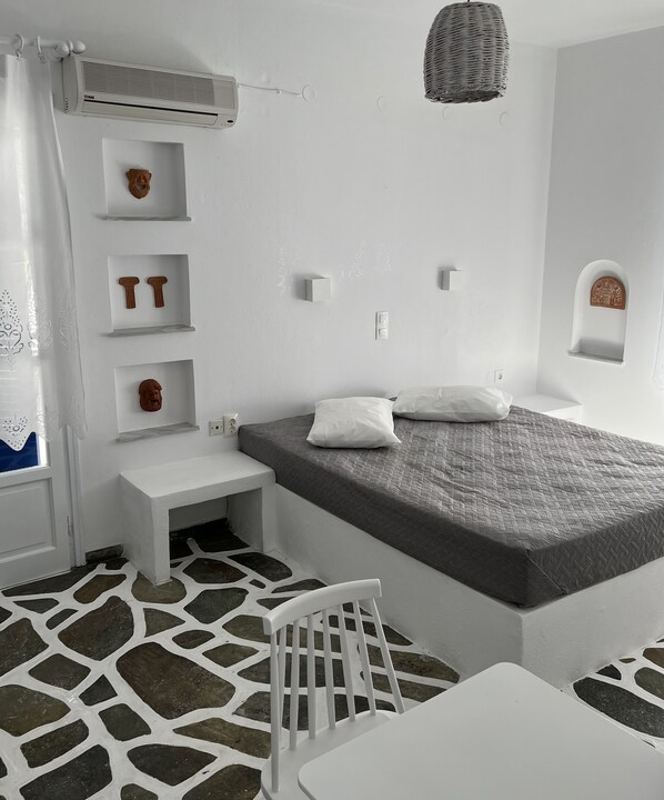 Una habitación minimalista con cama, una silla blanca, una mesita y objetos decorativos en estanterías.
