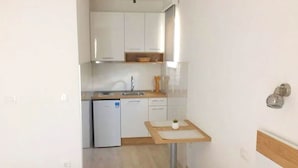 A compact kitchen with white cabinets, a small sink, and a dishwasher. There is a wooden table with a single chair and a small potted plant.