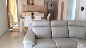 A modern living room with a grey sofa, a dining area with white chairs, and a kitchen with white cabinets and appliances.