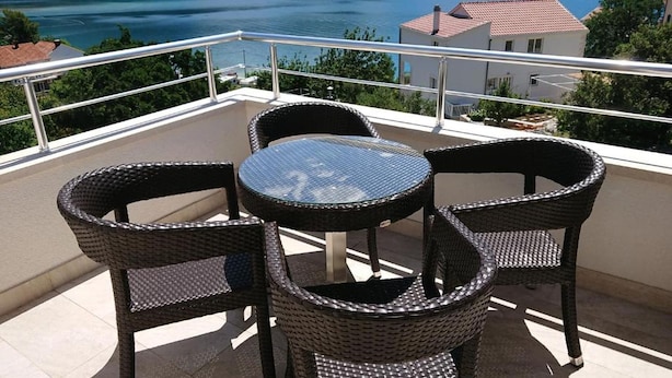 A balcony with wicker chairs and a glass table overlooking a lake with boats and a building in the background.