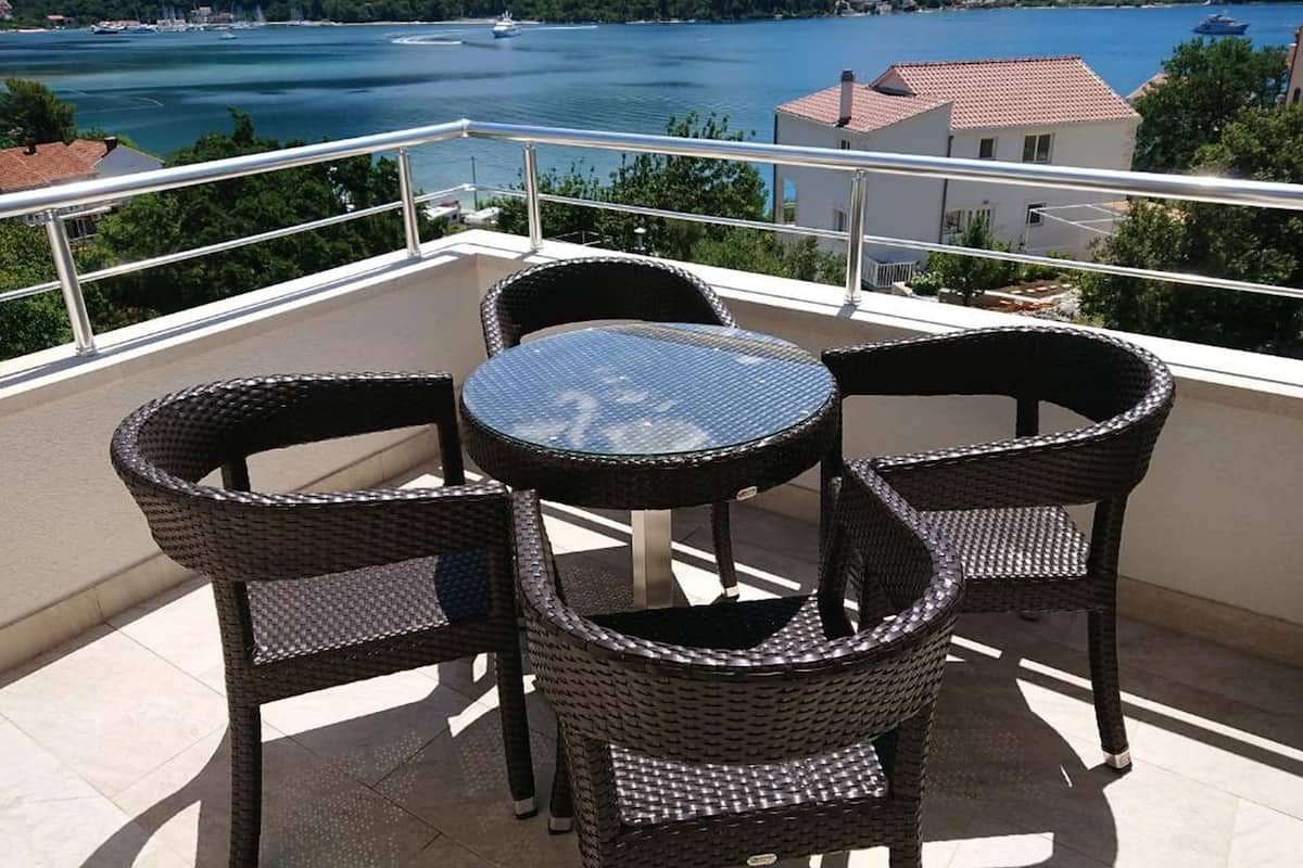 Un balcón con sillas de mimbre y una mesa de vidrio con vista a un lago con barcos y un edificio al fondo.