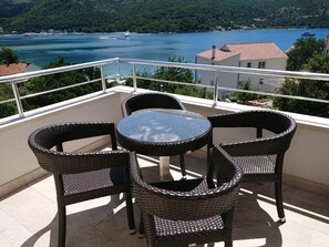 A balcony with wicker chairs and a glass table overlooking a lake with boats and a building in the background. - Sea Apartments Slano (Dubrovacko Primorje)