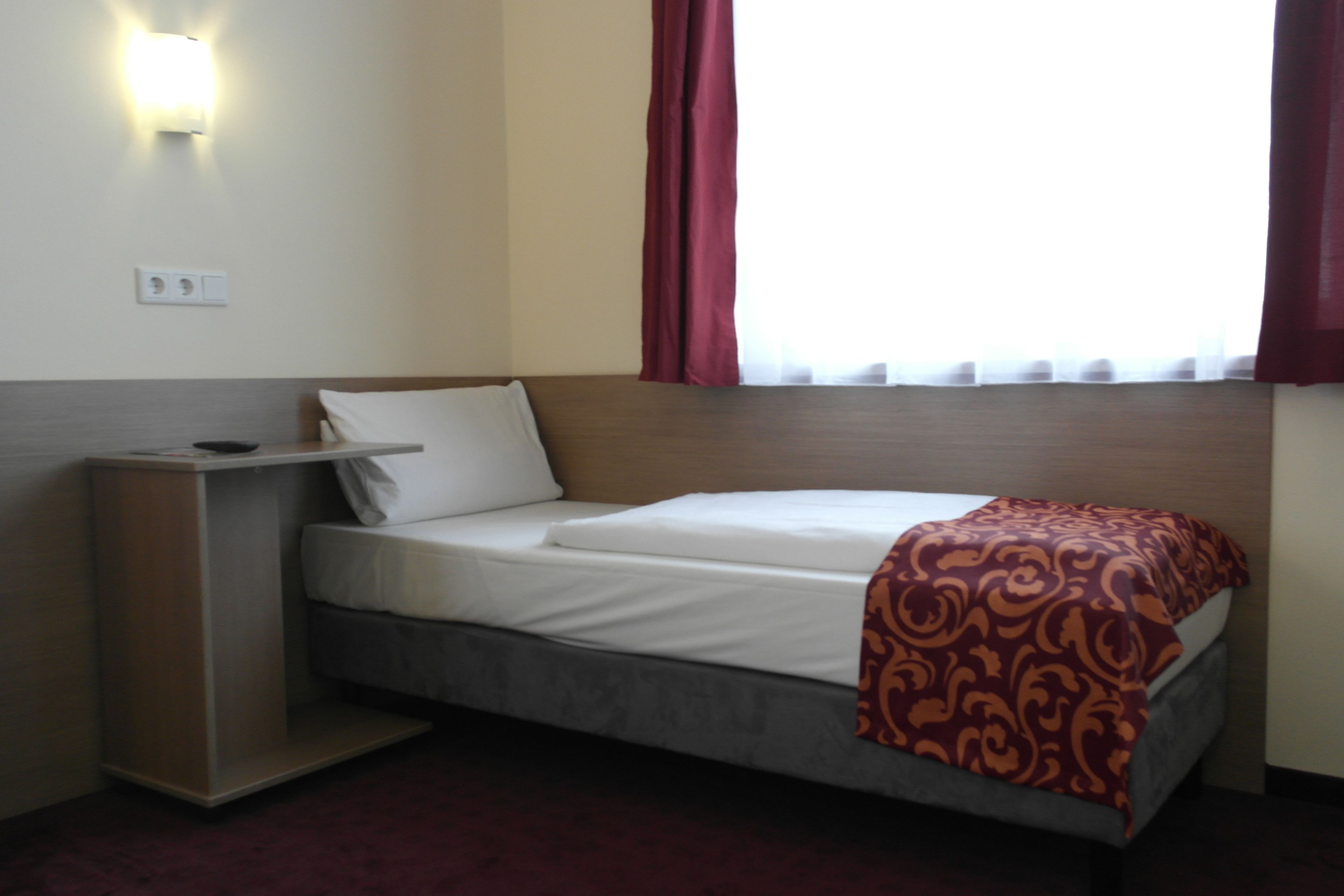 A hotel room with a bed, a bedside table, a wall-mounted light, and a window with curtains.