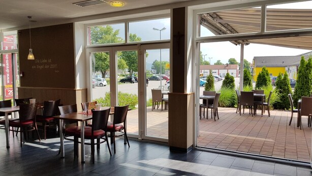 A restaurant with indoor and outdoor seating, large windows, and a view of the parking lot. - Dream Inn Hotel Regensburg (Tegernheim)
