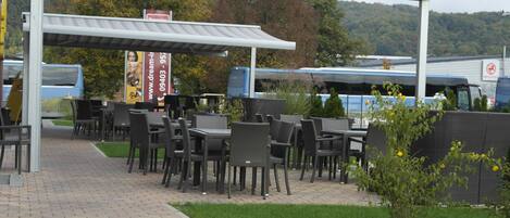 An outdoor seating area with tables and chairs, a covered walkway, and a view of a forested hill in the background.