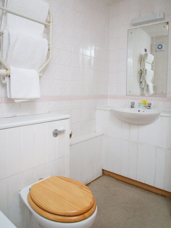 Un baño pequeño y limpio con azulejos blancos, tapa de madera para el inodoro, un lavabo blanco y un perchero con toallas blancas.