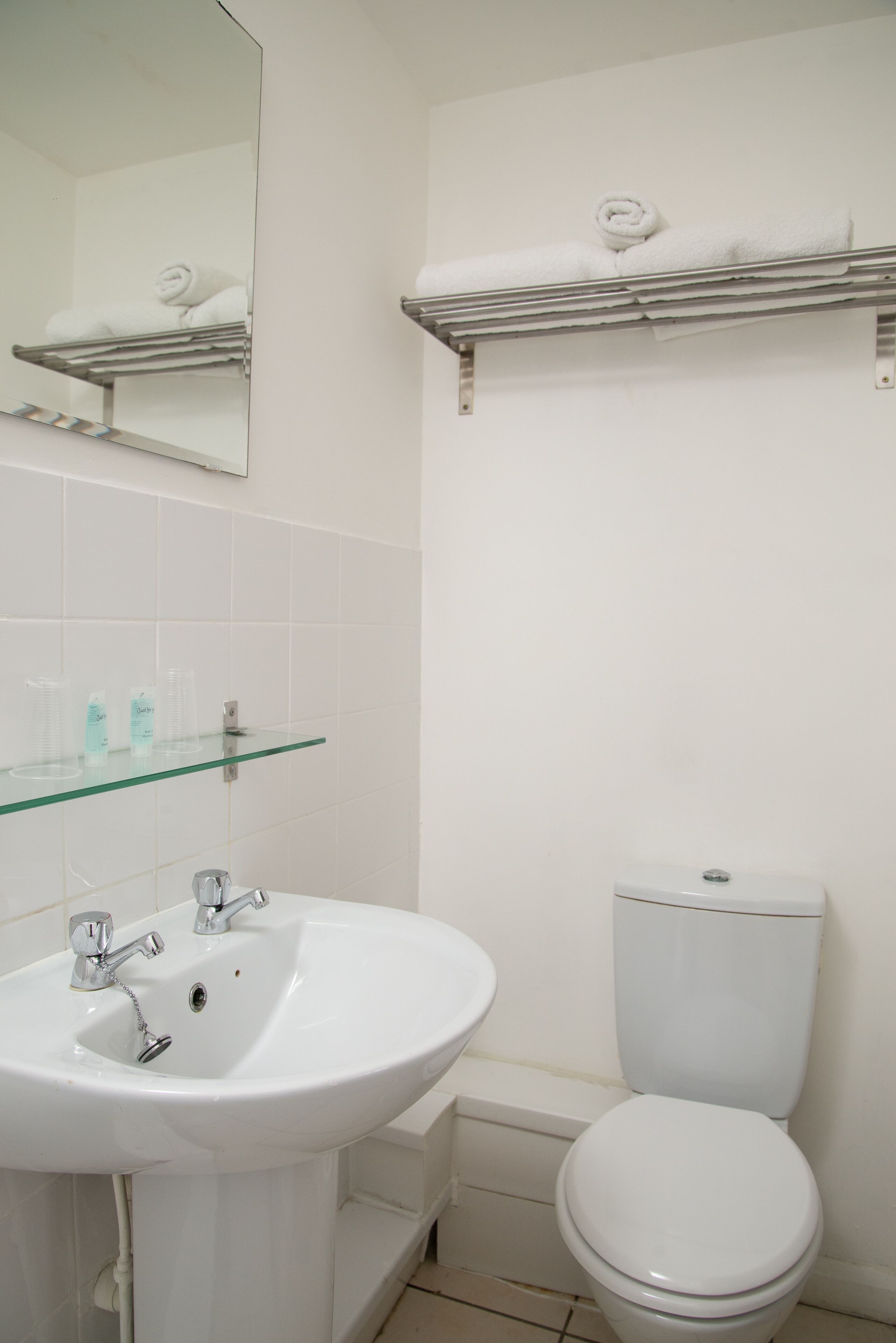 Een badkamer met een wit wastafel, spiegel en toilet. Boven het toilet zijn er planken met handdoeken.