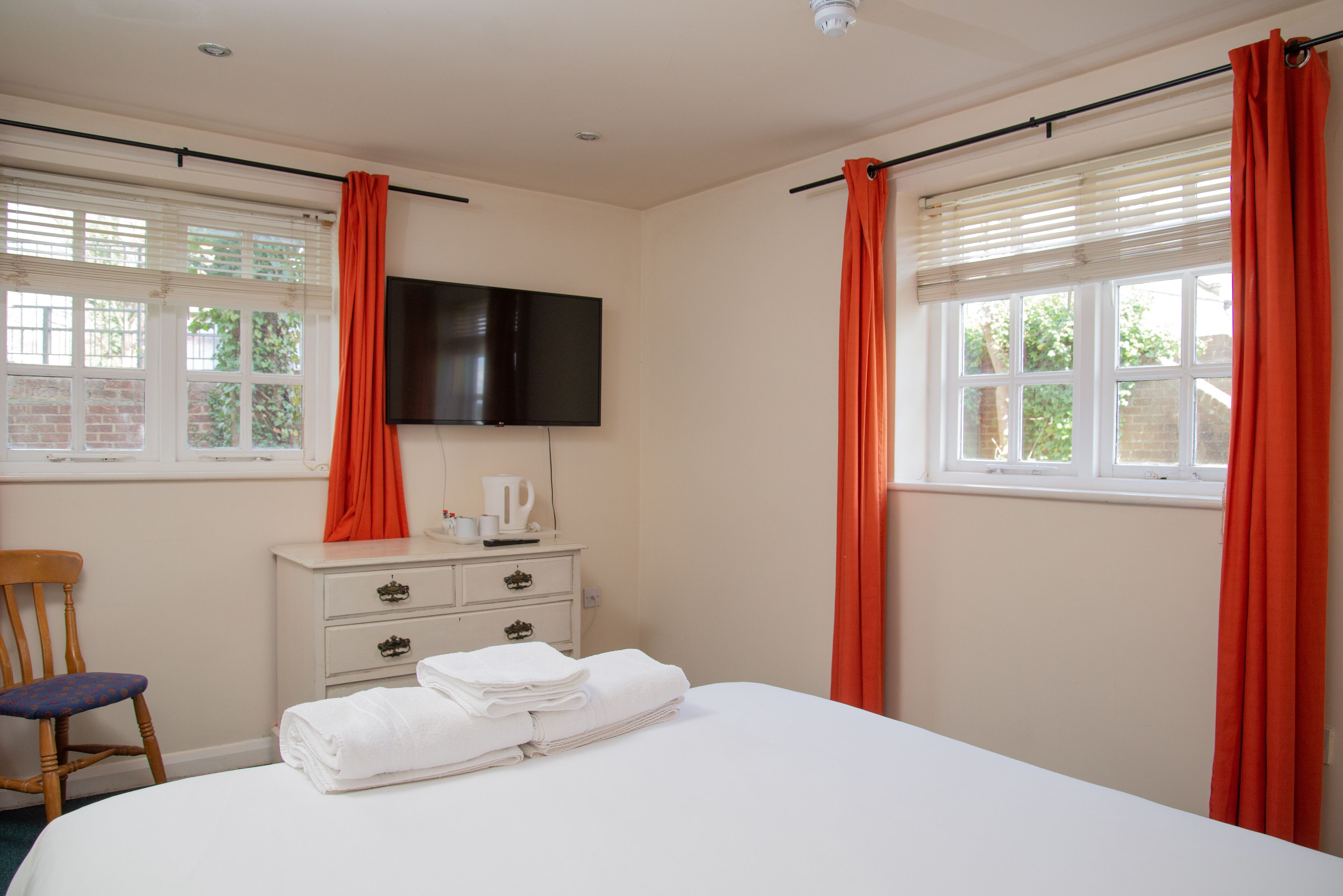 A hotel room with a bed, a television mounted on the wall, a bedside table with a kettle, and a window with curtains.
