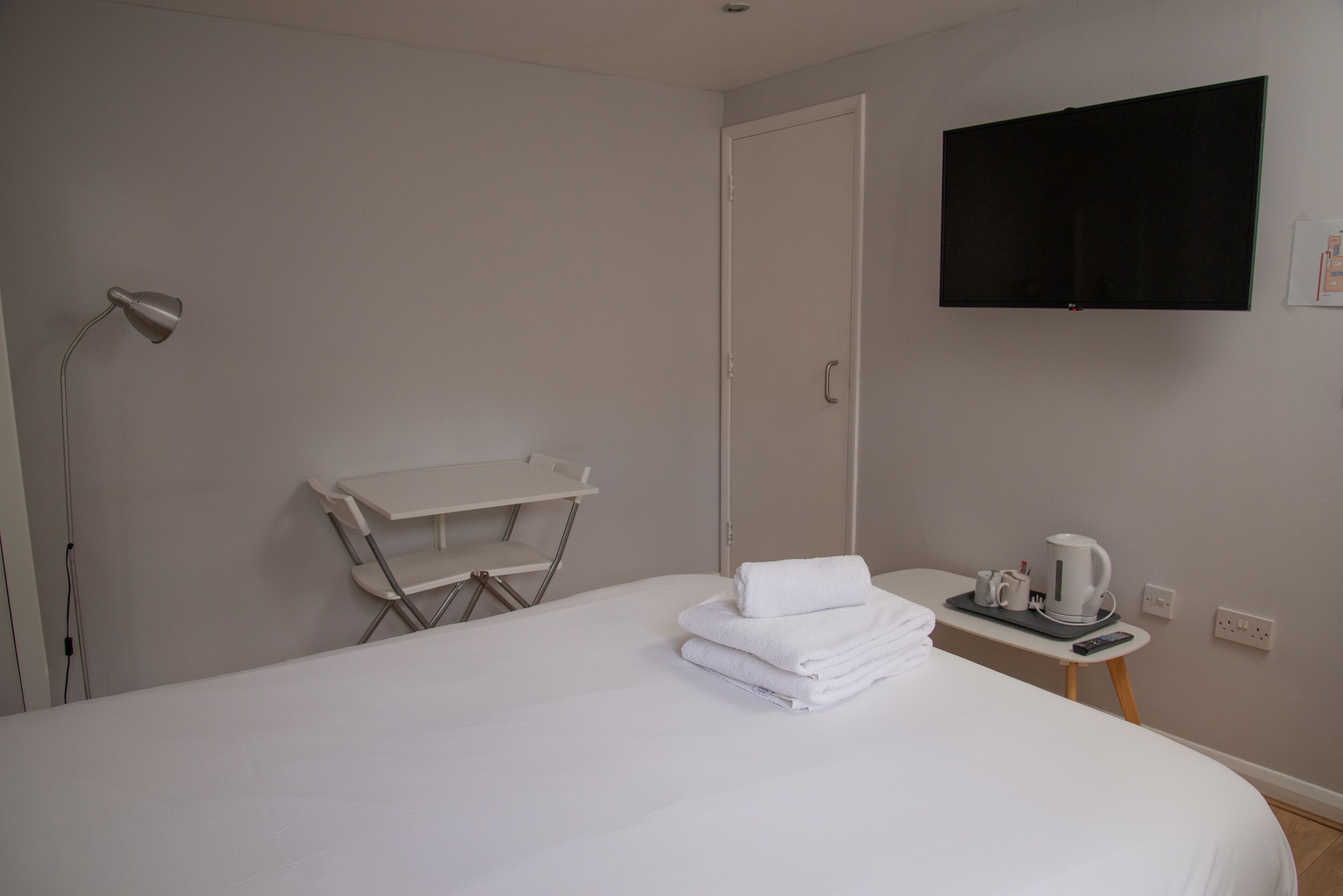 A hotel room with a bed, a desk with a chair, a flat-screen TV mounted on the wall, and a small table with a kettle and cups.