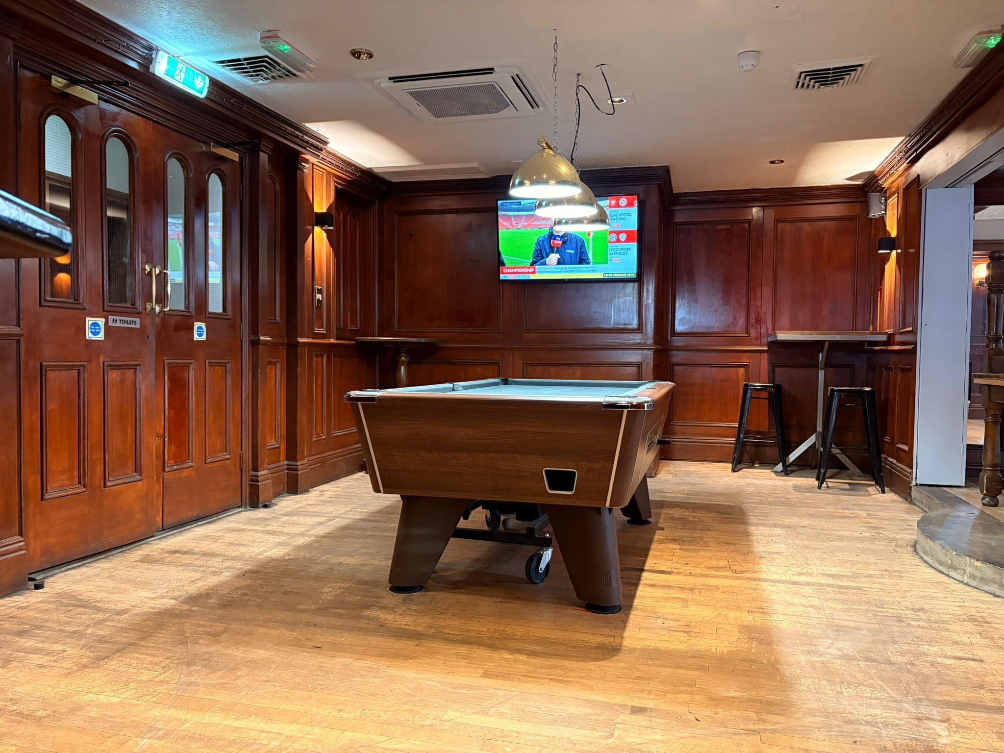 A billiards table in a room with wooden paneling and a television displaying a sports event.