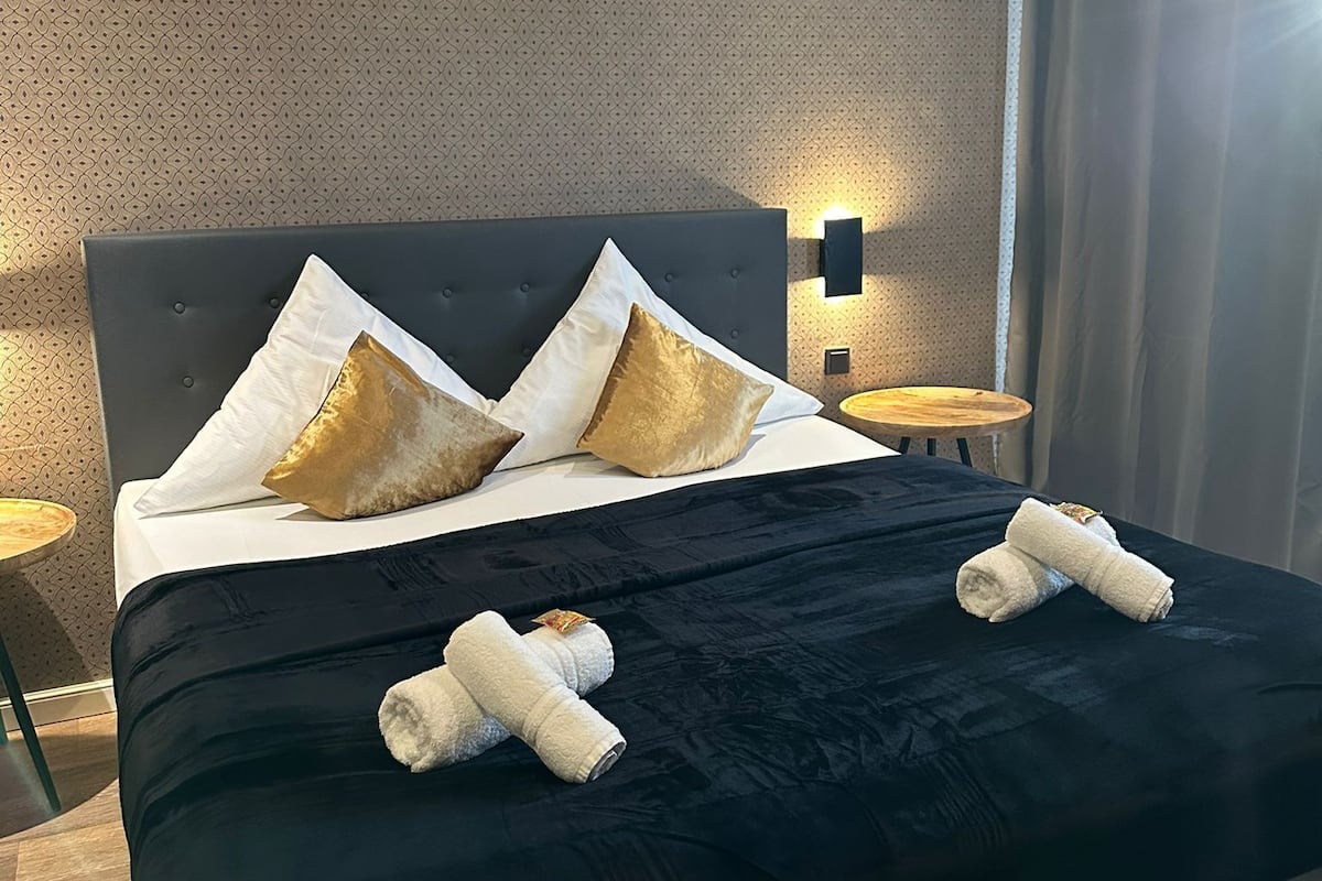 A neatly made bed with a black bedspread, two gold pillows, and two white towels.