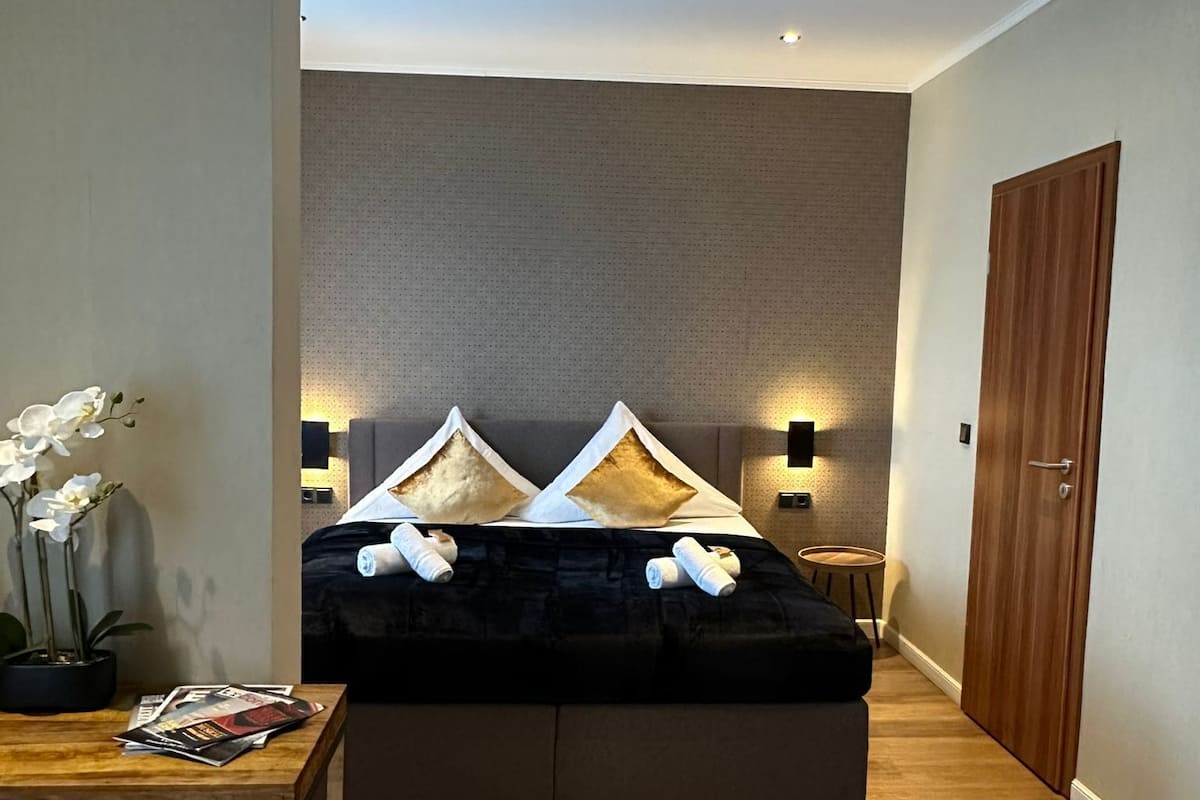 A modern hotel room with a large bed, a small table with magazines, a green chair, a wooden door, and a white rug.