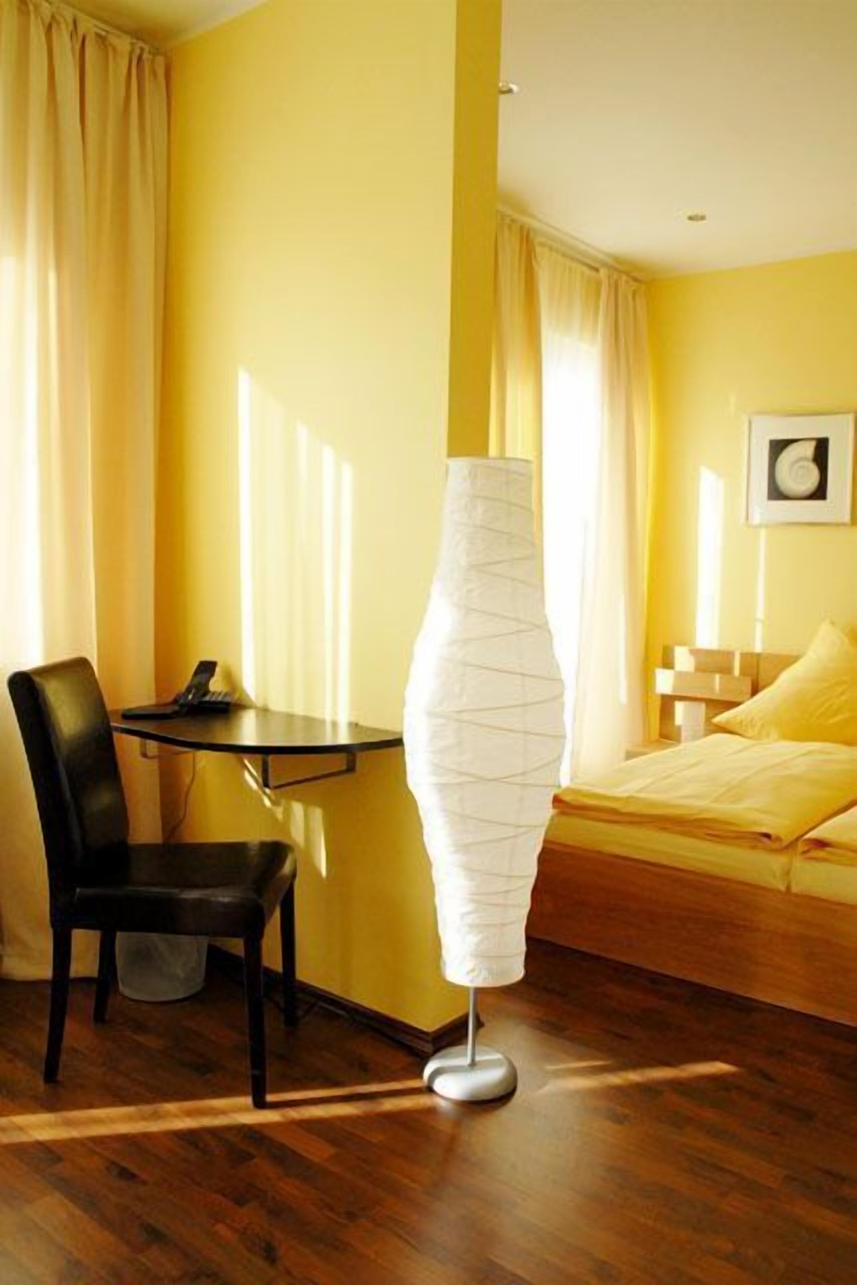 Una habitación de hotel con piso de madera, una cama con ropa de cama amarilla, un escritorio con silla y una lámpara de pie.